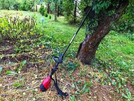 um cortador de grama fica perto de uma árvore. uma máquina de cortar grama fica perto da macieira, esperando o jardineiro. colhendo grama de outono amarelada. ao lado dos canteiros com terra e velhas plantas perenes foto