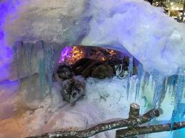 cena de design festivo um urso de brinquedo dorme em uma toca coberta de neve, uma árvore decorada atrás dele. pingentes de gelo decoram a entrada da toca foto