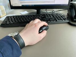 a mão de um homem com uma camisa e um relógio segura um mouse de computador na mesa da mesa do escritório com um computador com teclado. trabalho de negócios foto