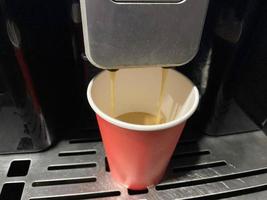 xícara de café de papelão vermelho na qual um delicioso café americano preto quente e revigorante é derramado da máquina de café no escritório pela manhã foto