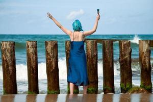 artista performática mulher de cabelos azuis em vestido azul em pé na praia segurando o pincel, vista traseira foto