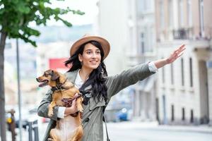 jovem mulher com cachorro acenando para parar um táxi na rua. jovem fêmea com cachorro engraçado esticando o braço e pegando táxi em pé na beira da estrada na cidade foto