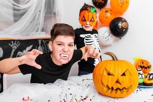 menino assustador com presas de vampiro olhando para a câmera na festa de halloween. jack o 'lantern abóbora de halloween em cima da mesa foto