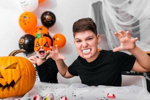 menino assustador com presas de vampiro olhando para a câmera na festa de halloween. jack o 'lantern abóbora de halloween em cima da mesa foto