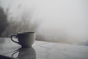 uma xícara de café branco descansa na beira de uma varanda de azulejos com uma espessa névoa branca ao fundo. foto