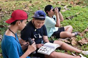 três meninos asiáticos estão lendo detalhes de pássaros e vão usar binóculos para observar pássaros nas árvores durante o acampamento de verão, ideia para aprender criaturas e animais selvagens fora da sala de aula. foto
