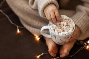 as mãos e os pés descalços da criança seguram a xícara de chocolate quente marshmallow fecham as luzes da guirlanda. conceito de natal, férias feliz ano novo menina sentada no sofá dentro foto