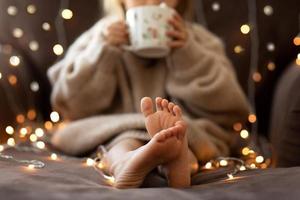 mãos e pés descalços de crianças fecham luzes de guirlanda. suéter bege de malha felpudo felpudo e quente. conceito de natal, feriado. feliz ano novo. foco seletivo. menina criança sentada no sofá dentro de vibrações aconchegantes foto