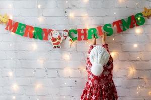 menina de chapéu de malha com pompom decorado quebra parede branca em casa guirlanda feliz natal e luzes foto