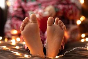 close-up criança pés descalços garoto de pijama vermelho de natal segurar caneca copo listrado verde vermelho doces bastões de natal bokeh luzes fundo. ano Novo foto