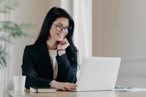 alegre funcionária digita no computador portátil, trabalha remotamente, concentrado com sorriso na tela do dispositivo, escreve carta de e-mail, trabalha no escritório bebe café usa novo aplicativo. conceito de tecnologia foto