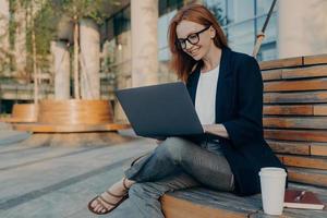 feliz e alegre mulher ruiva europeia trabalhando remotamente no laptop do lado de fora, sentado no banco de madeira foto