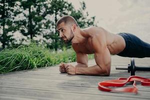 homem caucasiano barbudo fitness com torso nu fica em pose de prancha, exercícios ao ar livre, posa perto de equipamentos esportivos, tem braços fortes, demonstra resistência e motivação. treino abdominal. foto