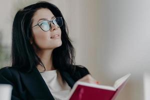 foto interna de jornalista feminina morena sonhadora pensativa trabalha na resenha do livro, segura o bloco de notas aberto, anota algumas ideias ou notas de memorando, cria um novo artigo, trabalha no projeto, usa óculos.