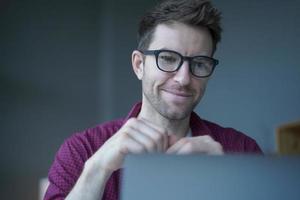 sorridente jovem alemão freelancer em óculos trabalhando remotamente on-line no laptop do escritório em casa foto