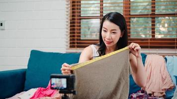 estilista asiático designer influenciador de moda mulheres usando streaming de câmera e ao vivo para vender roupas, produtos e acessórios, comércio eletrônico transmitido online no sofá na sala de estar em casa. foto