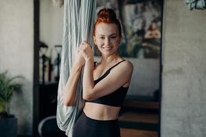 garota ruiva positiva em pé perto da rede sorrindo para a câmera esperando a aula de ioga aérea foto