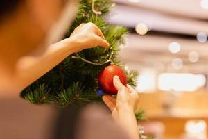 a mão de uma mulher pendura uma bola de natal na árvore de natal feriado festivo. foto