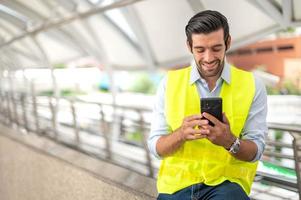 close-up homem caucasiano usa um smartphone para se comunicar com seu colega de trabalho e para contato com seu amigo enquanto ele usa colete amarelo e trabalha em seu site. foto