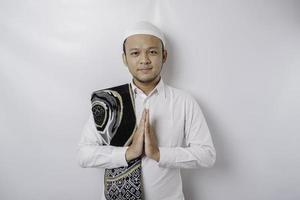 sorridente jovem muçulmano asiático com tapete de oração em seu ombro, gesticulando saudação tradicional isolada sobre fundo branco foto