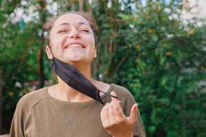 garota positiva feliz tira a máscara médica protetora do rosto ao ar livre. jovem removendo a máscara sorrindo após a vacinação. conceito covid 19 de pandemia de coronavírus. alergia ao pólen de flores de primavera. foto