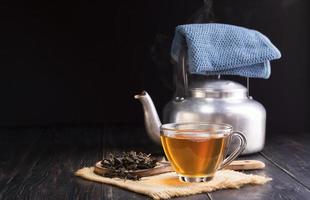 chá de ervas quente em uma xícara de vidro, xícara de chá e folhas de chá secas em colher de pau com chaleira colocada na mesa de madeira preta em fundo escuro. foto