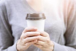 feche a mão feminina jovem segurando o copo de papel de levar embora bebendo café na luz solar natural da manhã. lugar de espaço para o seu texto ou logotipo. foto