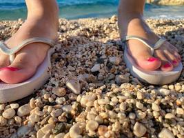 menina de pés com uma pedicure elegante em chinelos de praia elegantes femininos aqua no fundo da praia. duas sandálias na areia perto do mar. o conceito de férias na praia foto