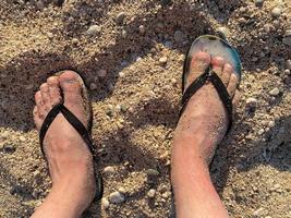 close-up do homem em pés de chinelos pretos em pé na praia, com uma onda de espuma suave abaixo deles. vista superior foto