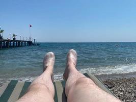 jovem descansa em uma espreguiçadeira de praia perto da piscina com água azul ao pôr do sol. férias de verão. pernas masculinas deitam-se numa espreguiçadeira perto do mar foto