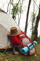 uma mochila com um tapete e um chapéu perto de uma barraca em uma grama em um acampamento em um fundo de floresta. foto