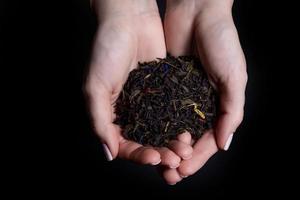 lindas mãos femininas seguram chá cru não fermentado e se oferecem para experimentá-lo. folha de chá seca na mão. chá com aditivos de bergamota isolados em preto foto