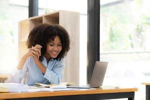 sorridente jovem empresária africana sentada em uma mesa em seu escritório em casa trabalhando on-line com um laptop foto