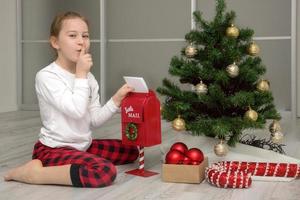 menina de pijama perto da árvore de natal joga uma carta para o papai noel na caixa de correio, presentes de natal foto