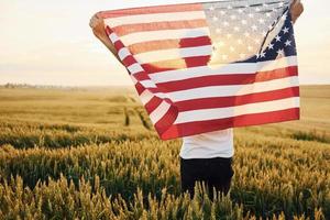 vista de trás. segurando a bandeira dos eua nas mãos. homem estiloso sênior patriótico com cabelos grisalhos e barba no campo agrícola foto