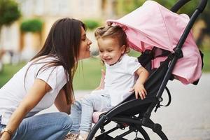 linda jovem mãe e sua filhinha no carrinho de bebê rosa andam na cidade foto