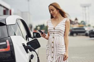 telefone nas mãos. mulher na estação de carga de carros elétricos durante o dia. veículo novo foto