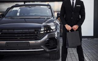 close-up vista do empresário de terno preto e gravata fica perto de automóvel moderno com sacolas de compras nas mãos foto
