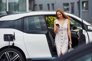 telefone nas mãos. mulher na estação de carga de carros elétricos durante o dia. veículo novo foto