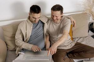 casal gay feliz com roupas casuais passando tempo juntos em casa e assistindo filme no laptop. dois homens caucasianos relaxantes. relações homossexuais e amor alternativo. interior acolhedor. foto