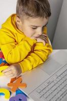 adorável, fofo, garotinho aprendendo alfabeto online, com laptop em casa. criança usando plataforma de e-learning para fazer uma videochamada com seu professor e estudar. educação domiciliar e educação a distância para crianças foto