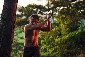 processo de cortar a árvore usando o machado. homem bonito sem camisa com tipo de corpo musculoso está na floresta durante o dia foto