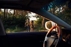 vista do interior do carro. volante, espelho lateral. lindo casal jovem se diverte na floresta durante o dia foto