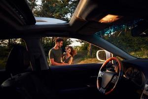 vista do interior do carro. volante, espelho lateral. lindo casal jovem se diverte na floresta durante o dia foto