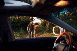 vista do interior do carro. volante, espelho lateral. lindo casal jovem se diverte na floresta durante o dia foto