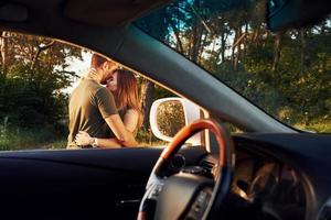 vista do interior do carro. volante, espelho lateral. lindo casal jovem se diverte na floresta durante o dia foto