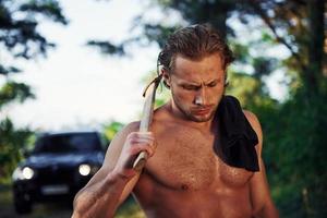 feche o retrato do lenhador com machado na mão. homem bonito sem camisa com tipo de corpo musculoso está na floresta durante o dia foto