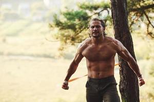 fazendo treino. homem bonito sem camisa com tipo de corpo musculoso está na floresta durante o dia foto