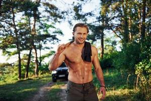 feche o retrato do lenhador com machado na mão. homem bonito sem camisa com tipo de corpo musculoso está na floresta durante o dia foto