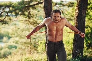 fazendo treino. homem bonito sem camisa com tipo de corpo musculoso está na floresta durante o dia foto